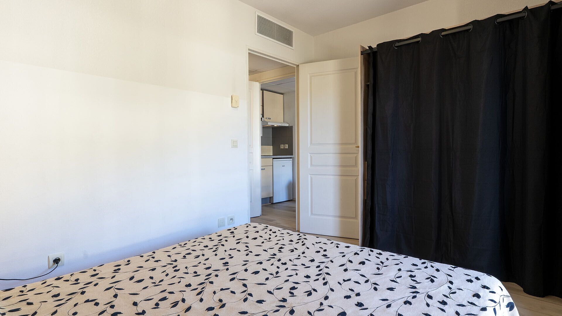 Un dormitorio con cama, cortina negra y una kitchenette al fondo.