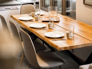 Une table à manger dressée avec des assiettes, des tasses et des couverts, des chaises, et une cuisine en arrière-plan.