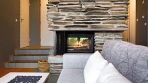A stone fireplace with a fire, a gray sofa, and a wooden table with black objects. - Rukatunturintie 8 g2 by Interhome (Kuusamo)