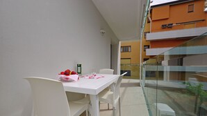 A white table with chairs, a glass partition, and a view of a building with balconies. - Comfortable Vacation Retreat With Balcony (Lignano Sabbiadoro)
