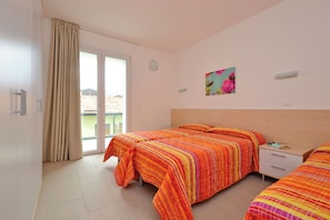 A hotel room with two beds, a sliding glass door to the balcony, a bedside table, and a wall-mounted light fixture. - Comfortable Vacation Retreat With Balcony (Lignano Sabbiadoro)