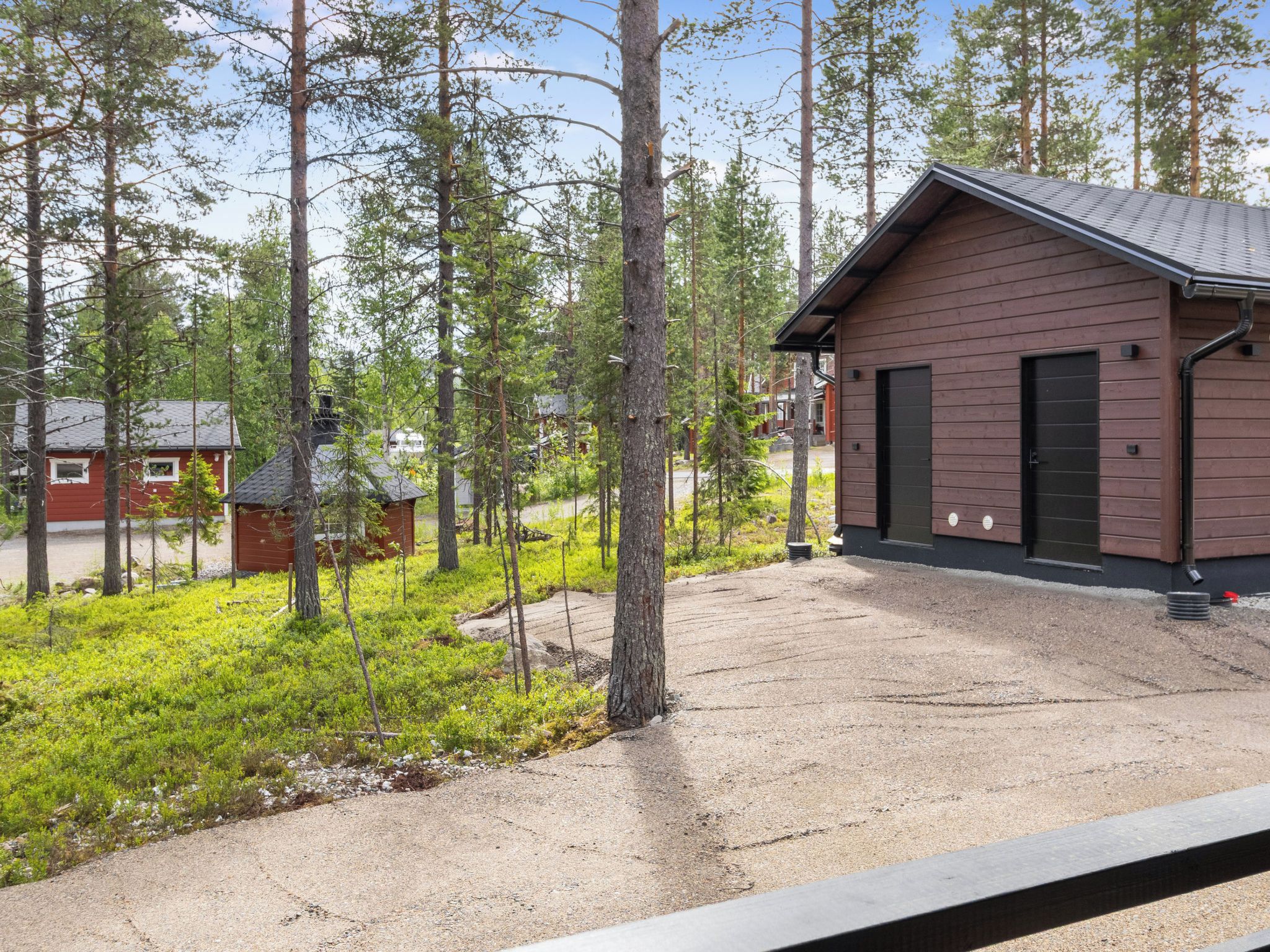 Mökki, jonka edessä on sorapäällysteinen ajotie, ja jonka ympärillä on puita. Taustalla näkyy pieni punainen rakennus.
