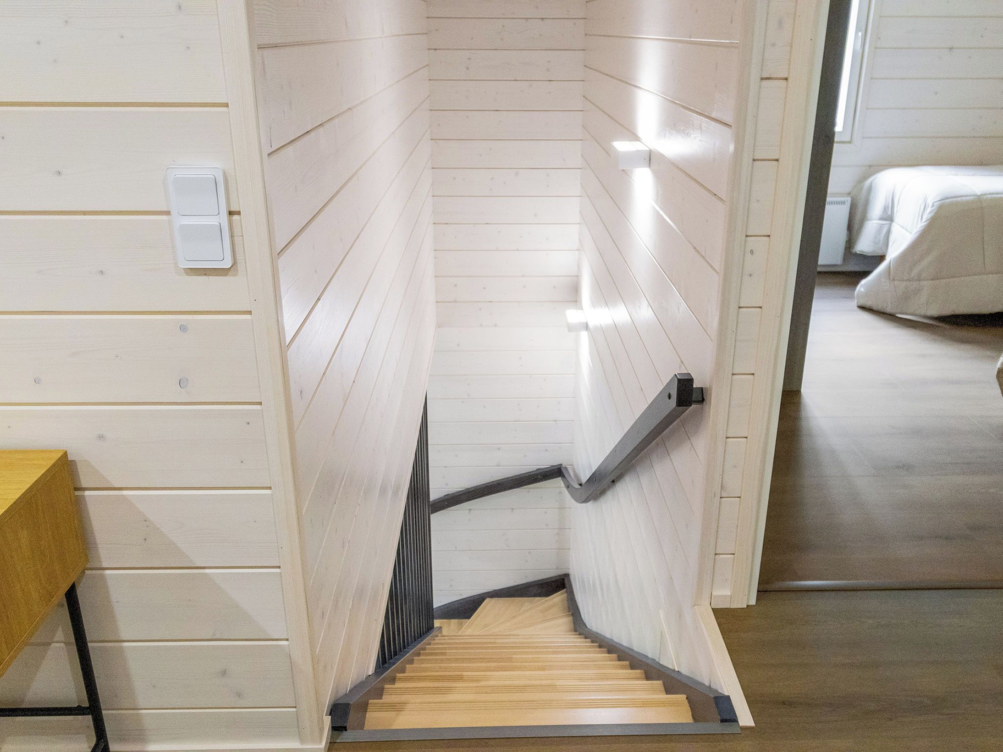 A wooden staircase with a handrail, leading to a bedroom with a bed.