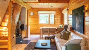 Interior de una cabaña de madera con escalera, comedor, sofá y un amplio ventanal.