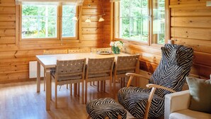 Interior de una cabaña de madera con una mesa de comedor, sillas, un sillón estampado con motivos de cebra y ventanas amplias con cortinas.