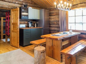 Interior rústico de una cabaña con mesa de madera, bancos, cocina con refrigerador y ventana con cortinas.