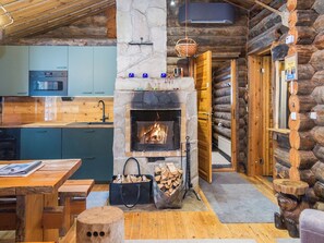 Interior de una cabaña de troncos con chimenea de piedra, una cocina con horno empotrado y una zona de comedor con mesa de madera.