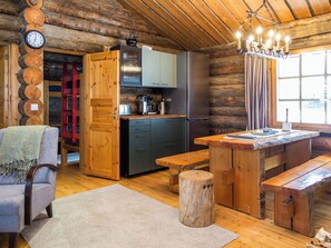 Interior de una cabaña de madera con una mesa de comedor de madera, bancos, una zona de cocina con electrodomésticos y un reloj en la pared.