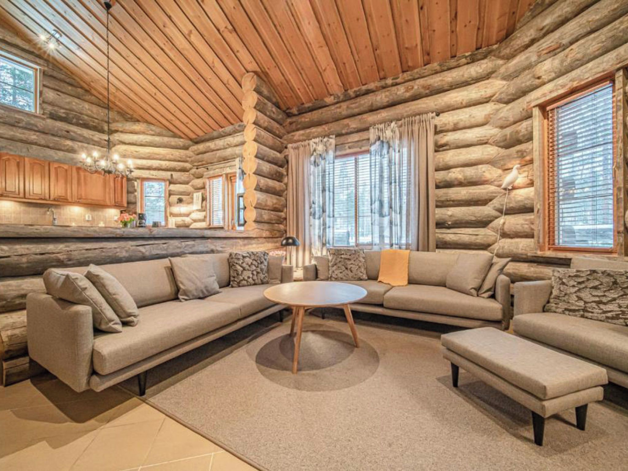 A log cabin interior with a large L-shaped sectional sofa, a round coffee table, and a kitchen area with wooden cabinets and a hanging light fixture.