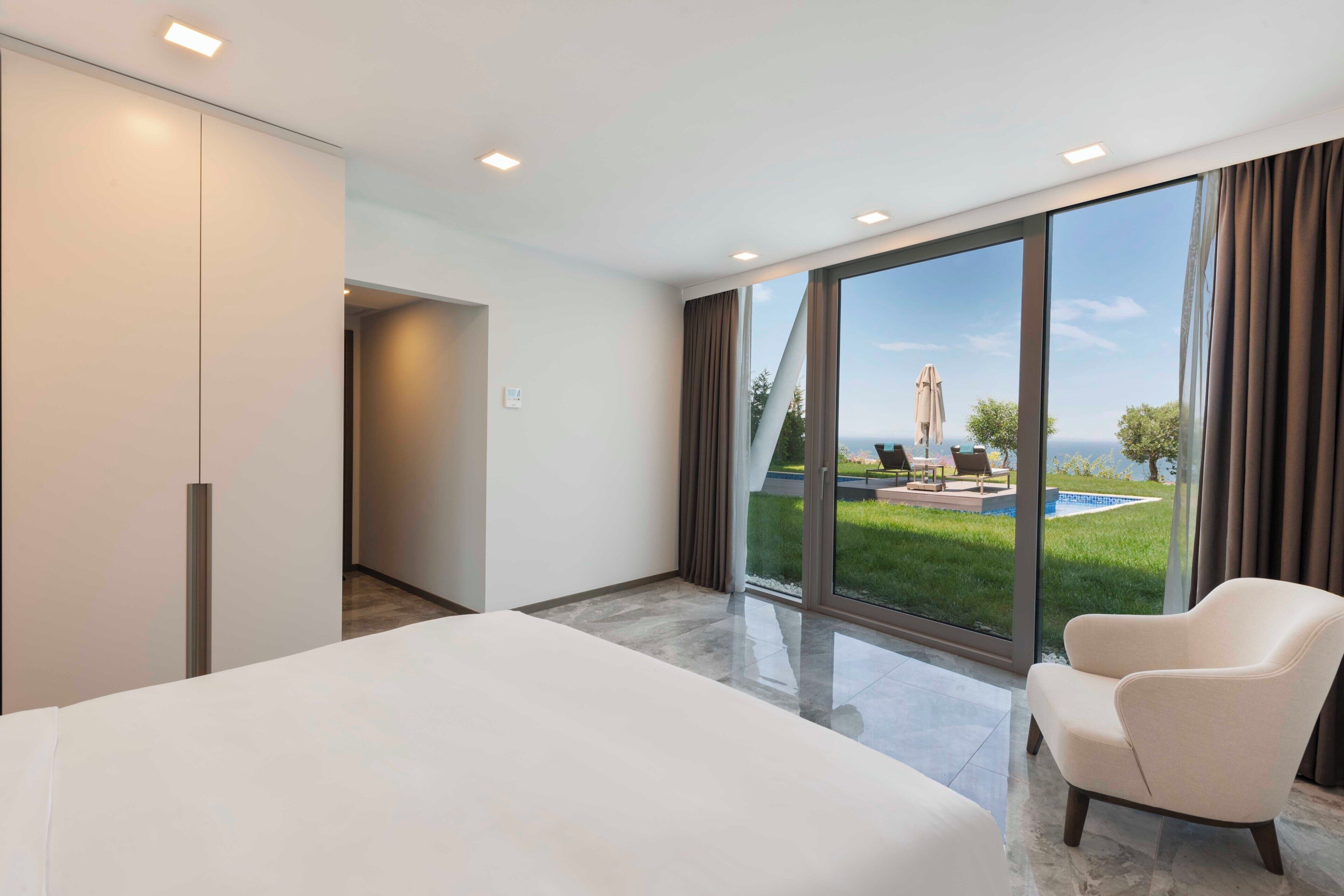 Una habitación de hotel moderna con una cama grande, una silla y vistas a una piscina y follaje.