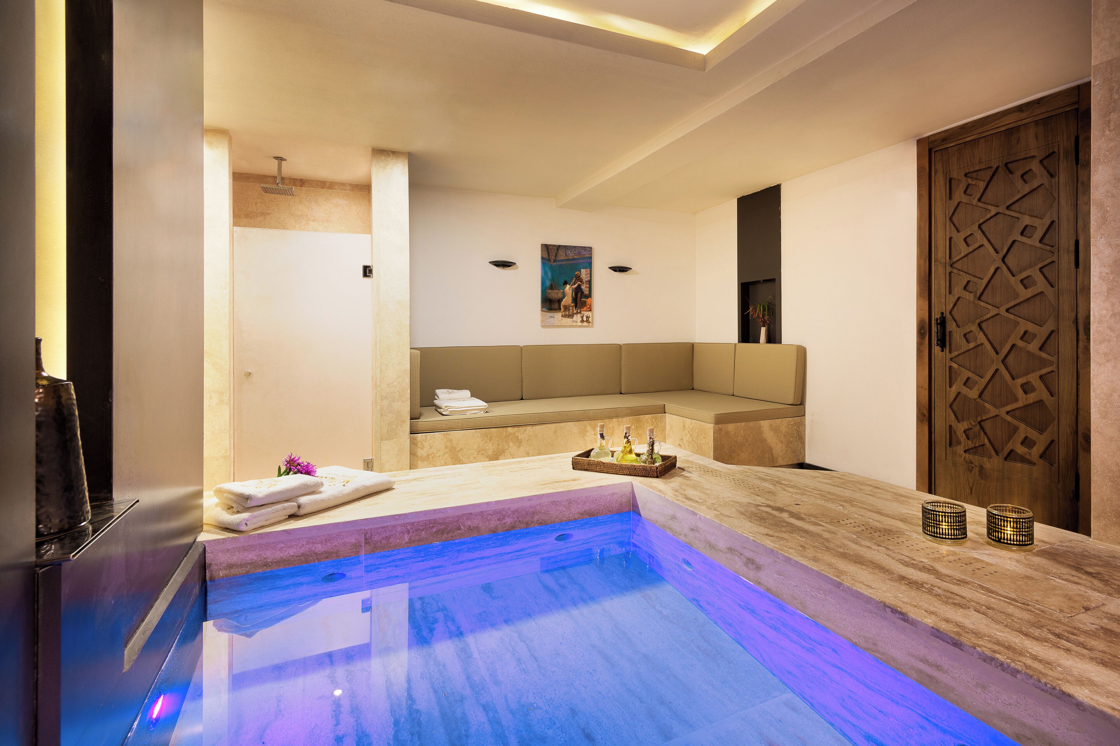 A modern spa area with a glass-enclosed hot tub, a seating area with a cushioned bench, and a wooden door with intricate carvings.