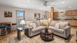 A living room with a sofa, coffee table, and bar stools. There is a kitchen in the background with wooden cabinets and a window. - Cardinals Nest In The Smokies ~ Family Friendly (Haywood County)