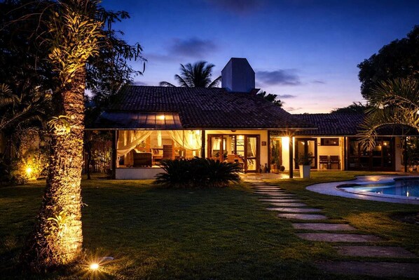 A house with a tiled roof, a swimming pool, and a well-lit garden at dusk.