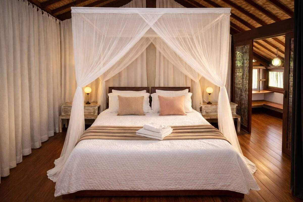 A four-poster canopy bed with white linens and two pillows, wooden flooring, and bedside lamps.