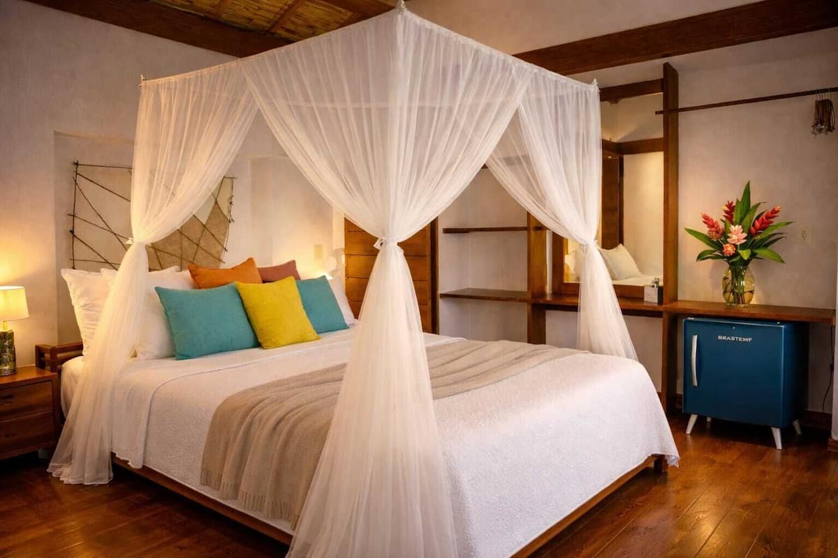 A four-poster canopy bed with a wooden headboard, a nightstand with a lamp, and a small blue cabinet with a flower vase.