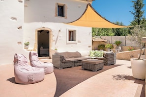 Eine Terrasse mit Gartenmöbeln, darunter eine Couch und Stühle, neben einem weißen Gebäude mit Tür und Fenstern.