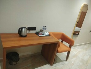 A small hotel room with a wooden desk, a chair, a coffee maker, a small trash bin, and a mirror on the wall. - Shreeja Central (Bhawanipatna)