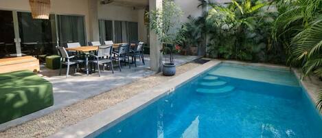 A swimming pool with a patio area, including a table and chairs, surrounded by greenery.