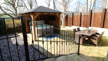 A wooden gazebo with a hot tub, a patio table with chairs, and a stone-tiled patio.