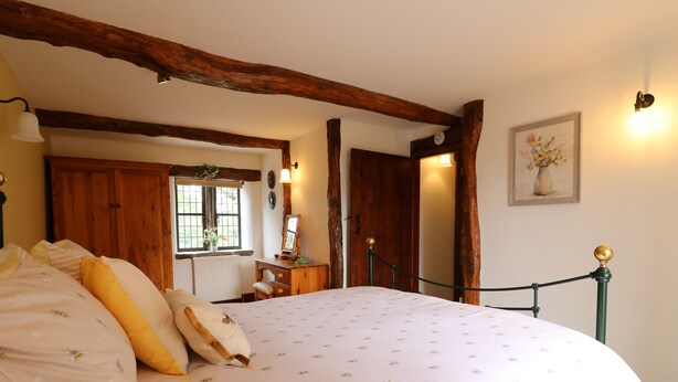 Een slaapkamer met een groot bed, houten balken, een dressoir, een spiegel en een raam met bloemen.