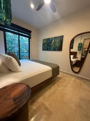 Un dormitorio con cama, ventilador de techo, ventana grande, un cuadro y un espejo.