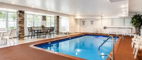 A swimming pool with a stone pillar, a table with chairs, and a white chair near the pool.