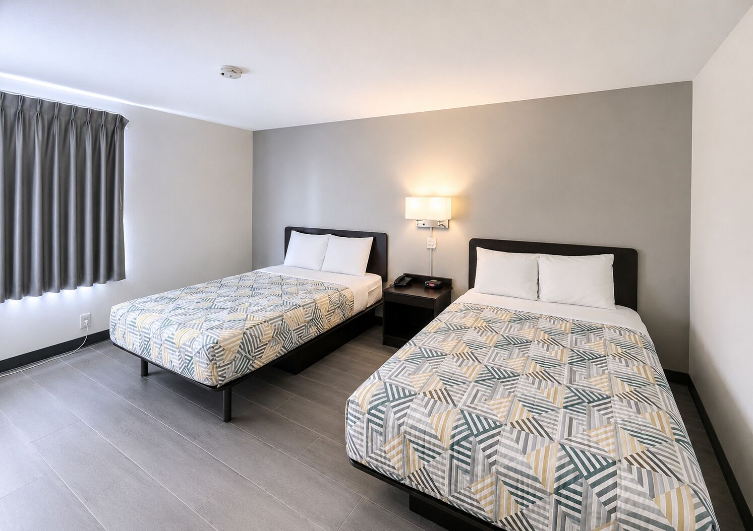 Une chambre d’hôtel avec deux lits, une table de chevet avec une lampe, une prise de courant et une fenêtre avec des rideaux.