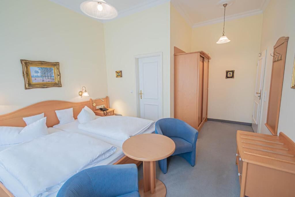 Habitación de hotel con cama doble, una mesita redonda, dos sillas azules, un armario y una lámpara.