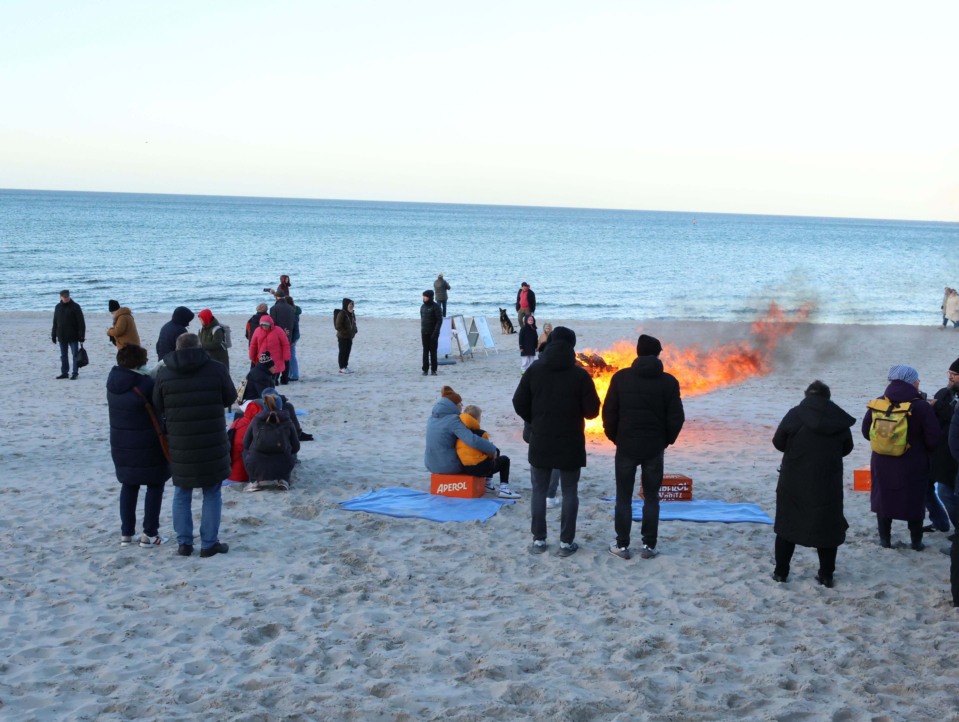 A beach scene with people gathered around a fire, some sitting on mats, and a few standing.