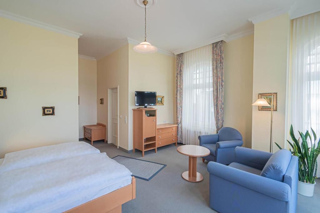 A hotel room with a bed, a desk, a television, a chair, a small table, and a potted plant.