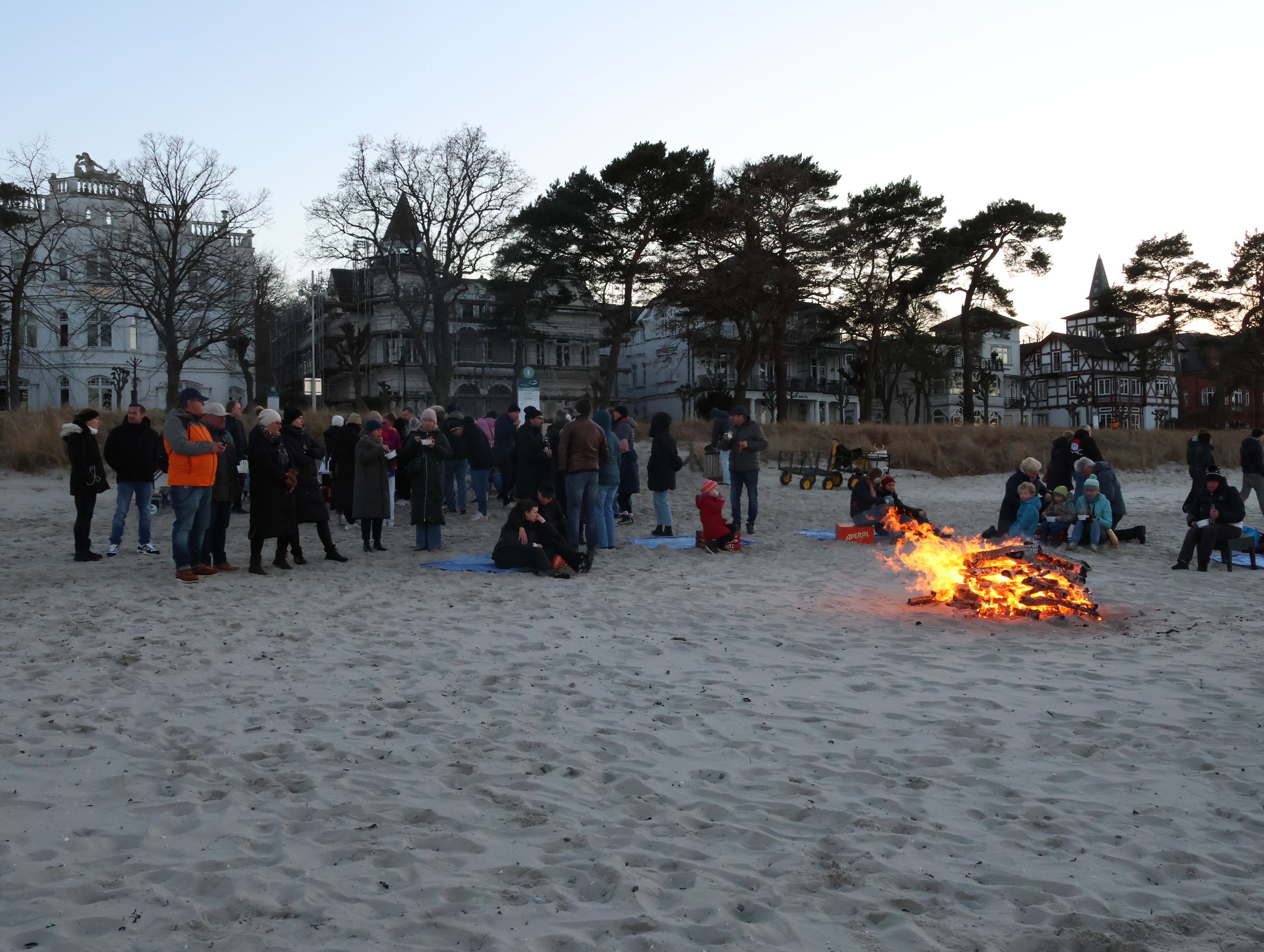 A sandy beach with people gathered around a fire, some sitting and others standing, with buildings in the background.