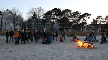 Una spiaggia sabbiosa con persone riunite attorno a un fuoco, alcune sedute e altre in piedi, con edifici sullo sfondo.