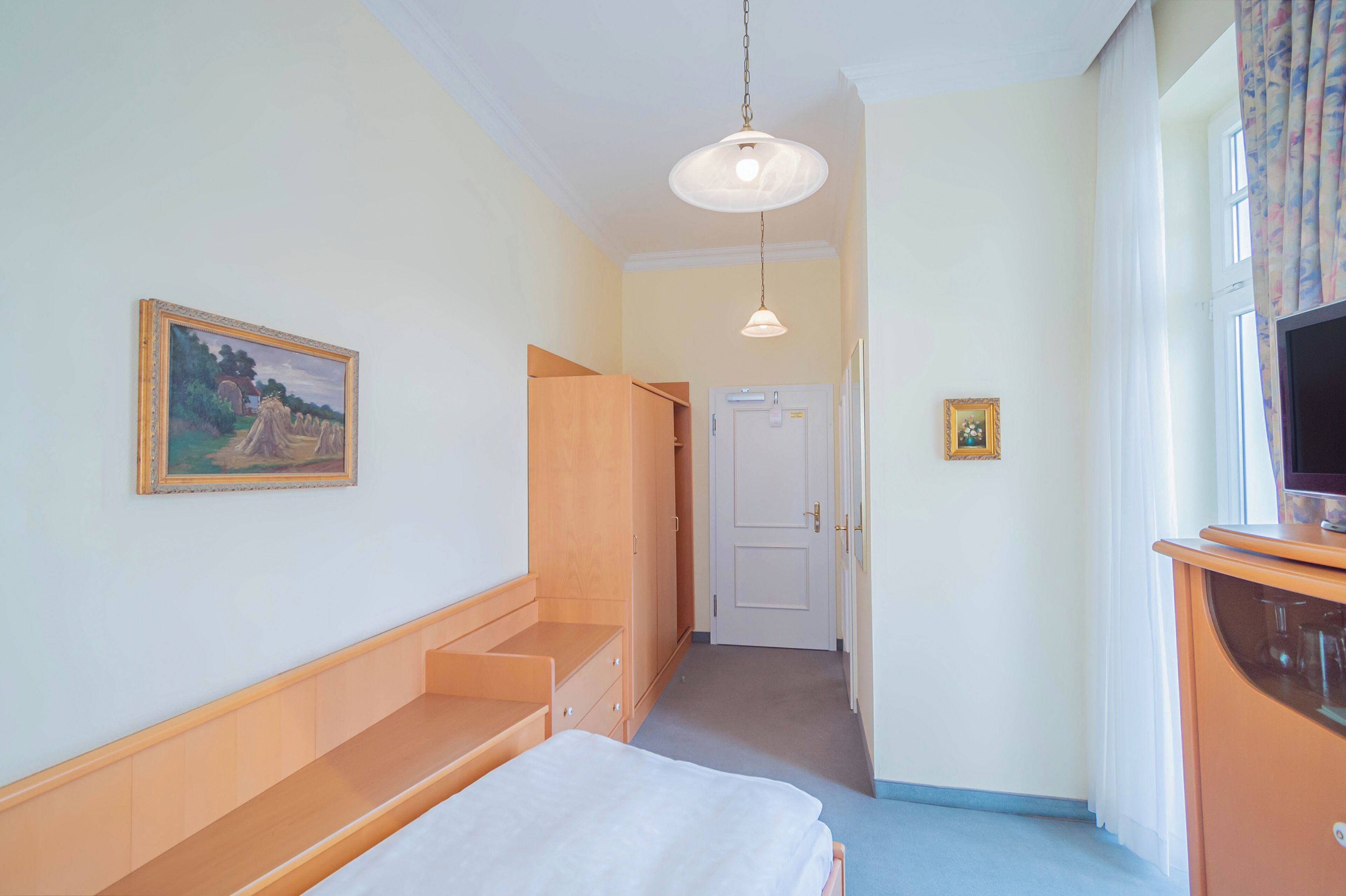 Un dormitorio con cama, armario de madera, un cuadro en la pared, televisor sobre un mueble bajo y una puerta que da a otra habitación.