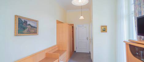 Un dormitorio con cama, armario de madera, un cuadro en la pared, televisor sobre un mueble bajo y una puerta que da a otra habitación.