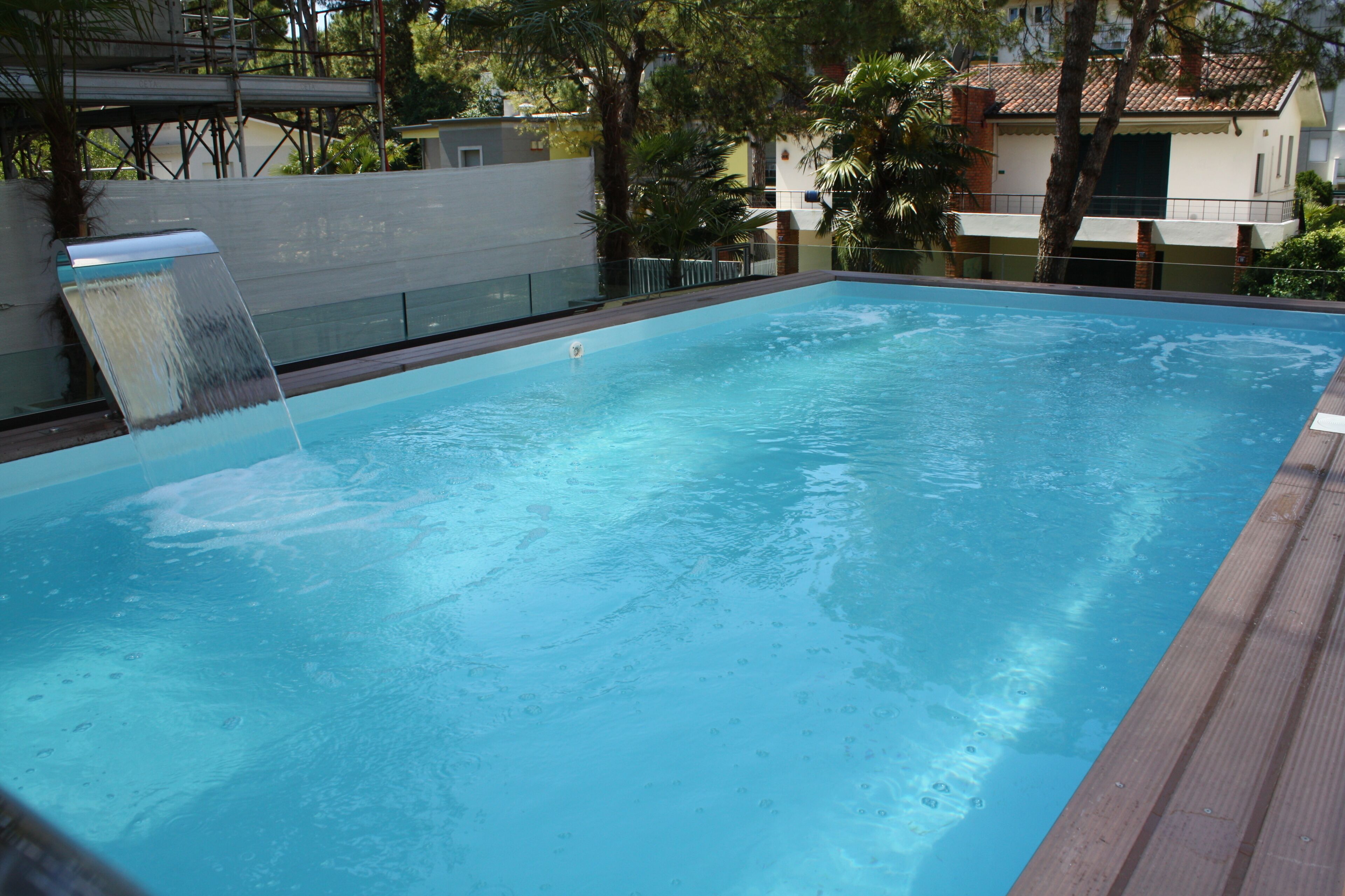 A rectangular swimming pool with a waterfall feature, surrounded by a wooden deck and a white wall.