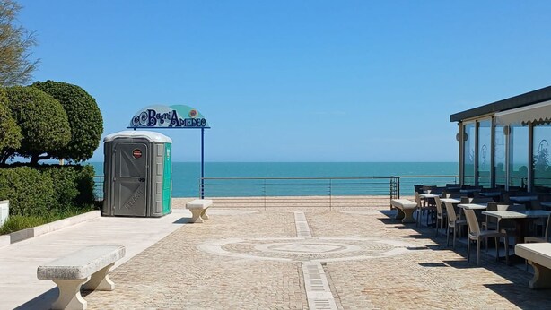 Un restaurante frente a la playa con mesas al aire libre, un baño químico y un letrero que dice 'Beoci Arma'.