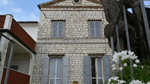 Un edificio de piedra con dos ventanas y contraventanas, un pequeño jardín y un árbol.