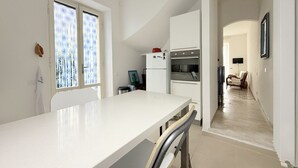 Una cocina con encimera blanca, un refrigerador y un horno. Hay una ventana con persianas, una silla y una puerta que da a otra habitación.