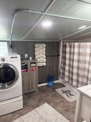 A laundry room with a washing machine, a drying rack, a blue trash bin, and a checkered shower curtain.