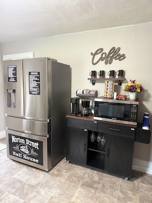 A kitchenette with a stainless steel refrigerator, a microwave, a coffee station, and a wall-mounted shelf with coffee mugs.
