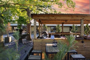 Un área de estar al aire libre con mesas y bancos de madera, rodeada de árboles y un edificio con techo de metal corrugado.