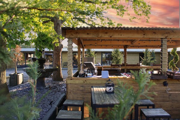 A wooden outdoor seating area with tables and benches, surrounded by trees and a building with a corrugated metal roof.