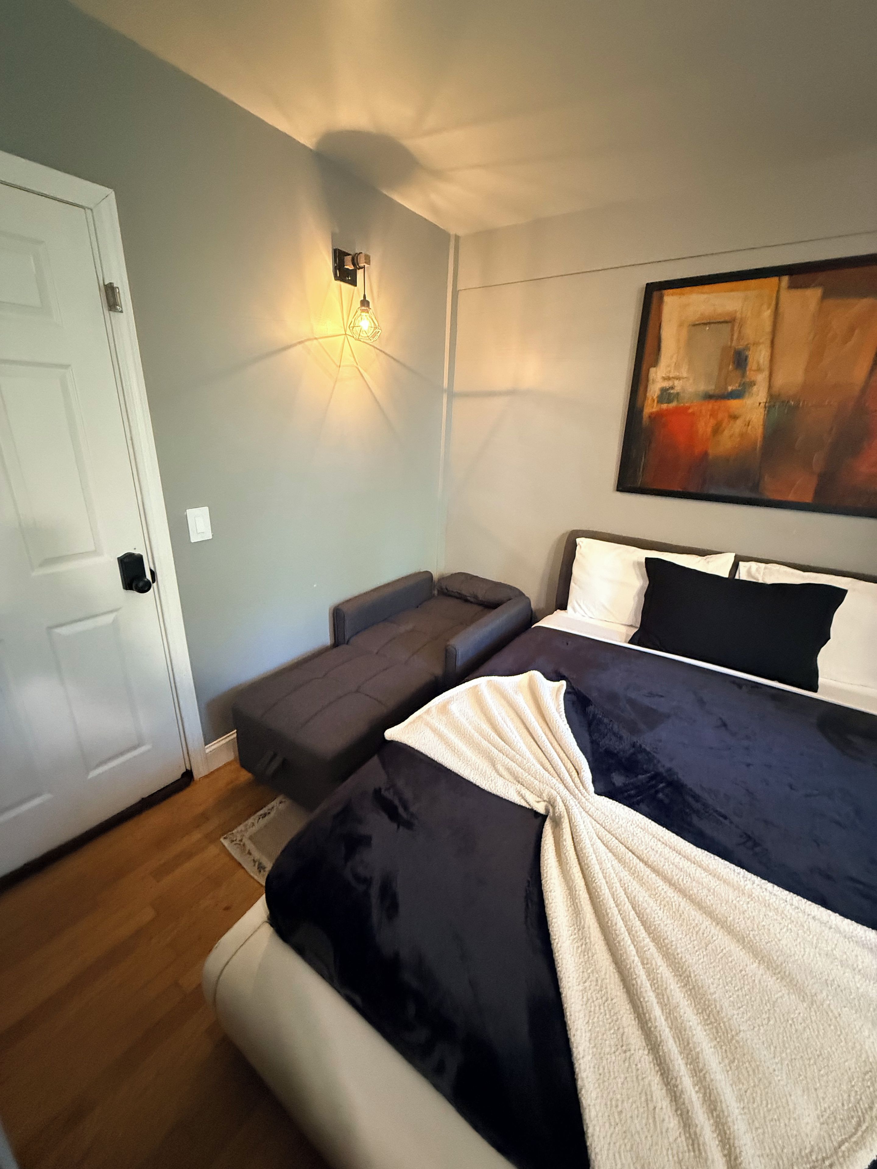 A bedroom with a bed, a sofa, a wall-mounted light, and a painting.