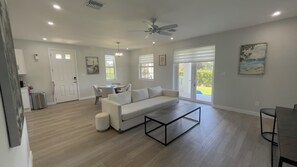 A spacious living room with a white sofa, a coffee table, and a dining area with a table and chairs. There is a ceiling fan, a fire extinguisher, and a window with blinds. - The Oasis Villa (Nassau)