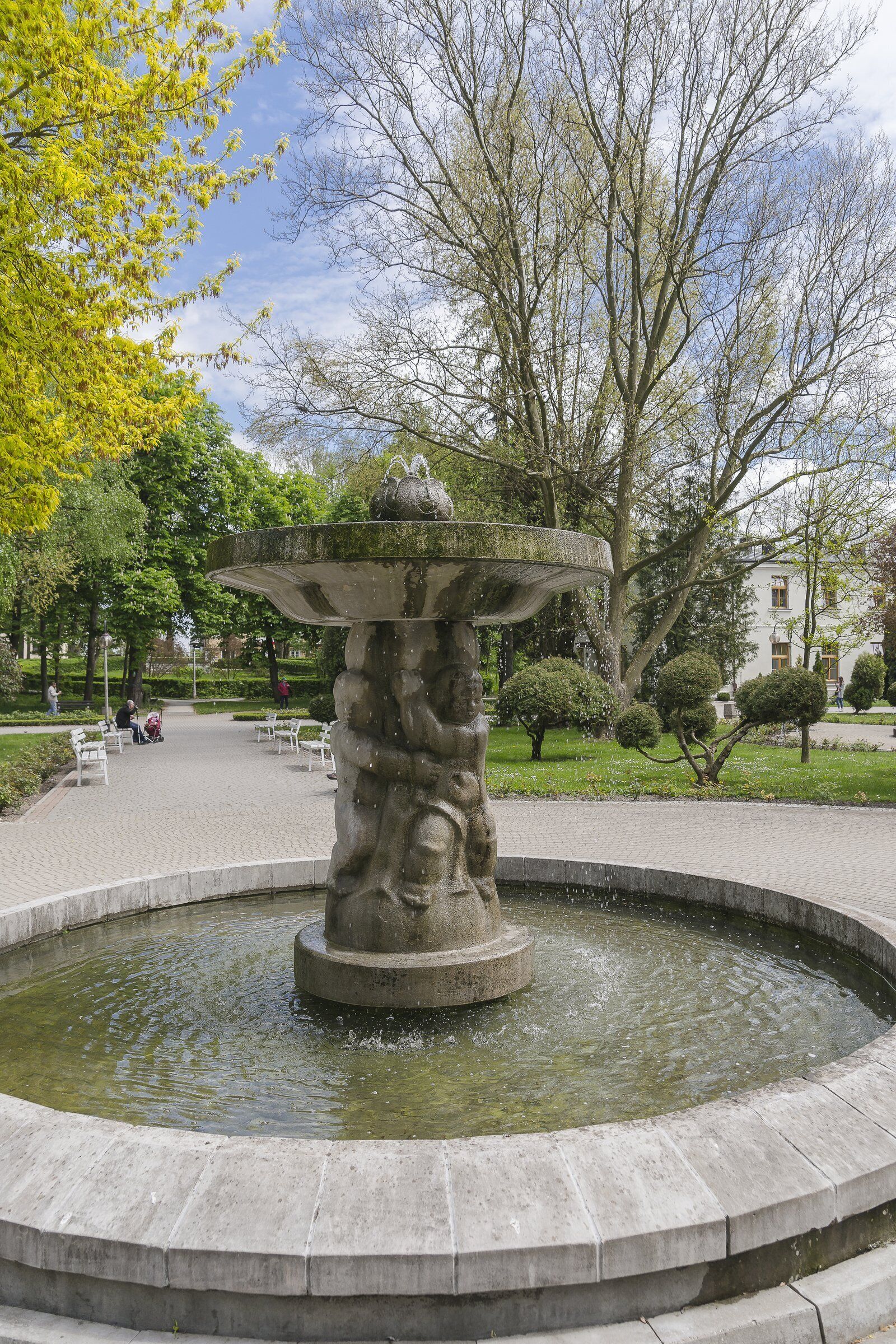 A fountain with a stone base and water feature, surrounded by a paved area with benches and trees.