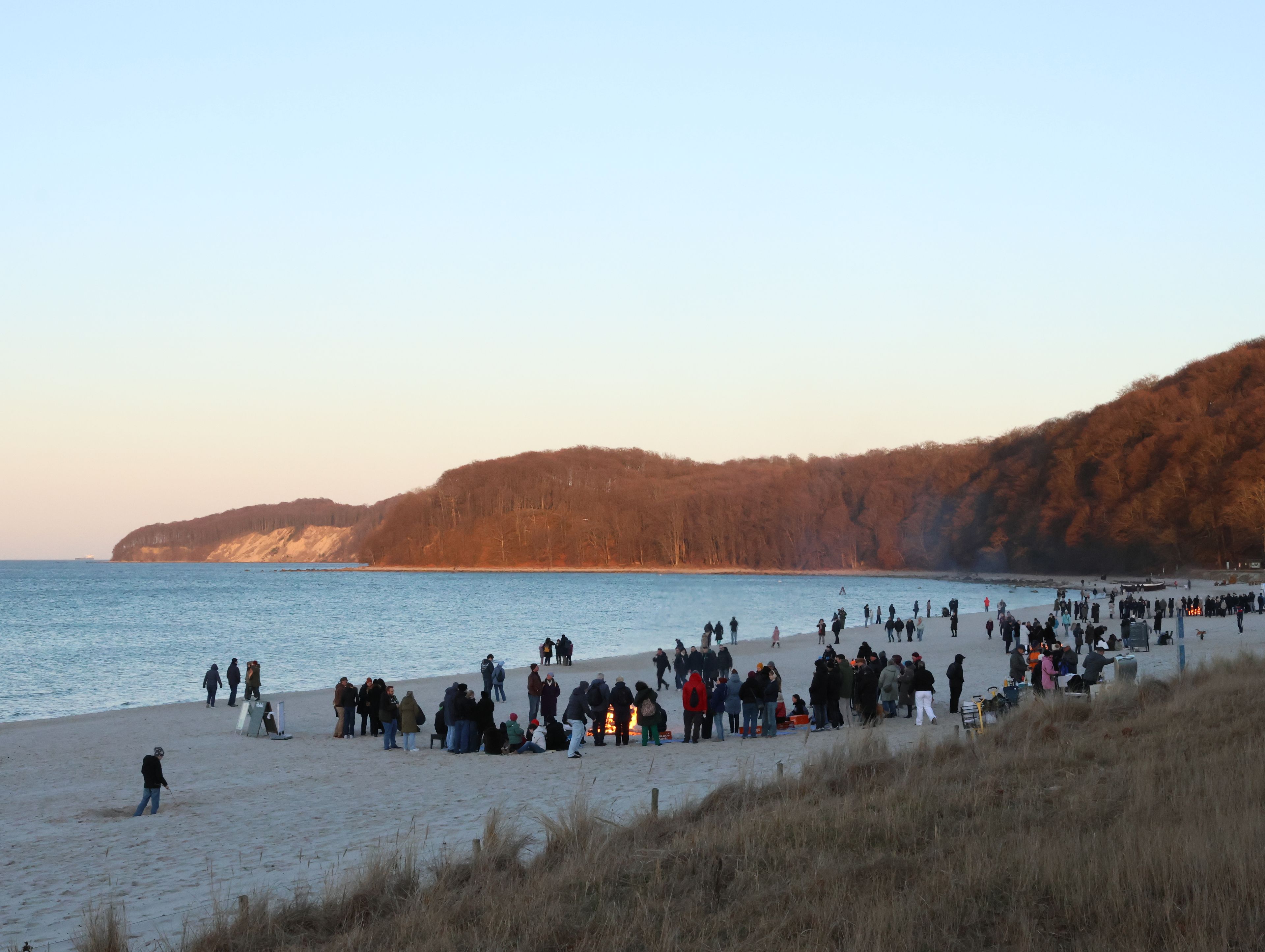 En strand med folk samlade, en sandstrand och en skogbeklädd kulle i bakgrunden.