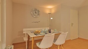 Un comedor con una mesa y sillas blancas, un reloj de pared y una mesita con una cesta y tetera.