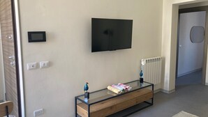 A modern living room with a flat-screen TV mounted on the wall, a glass coffee table with a wooden base, a radiator, and a door with a circular handle. - Newly renovated house, bright and within walking distance of Ortigia and the Greek theater. (Siracusa)
