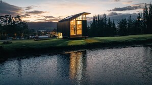 Une cabane moderne à la façade vitrée, située au bord d’un plan d’eau, entourée d’arbres et d’un paysage vallonné.
