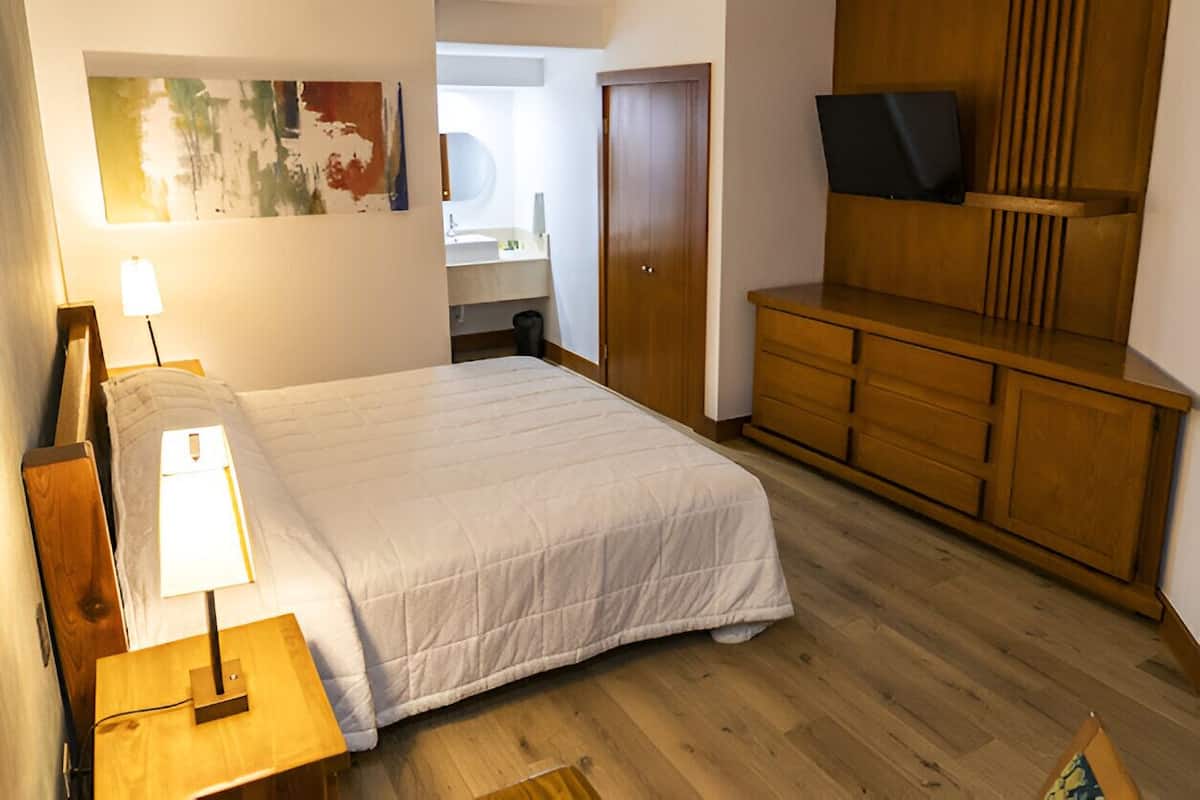 Un dormitorio con cama, mesita de noche, lámpara, cómoda de madera, televisor y un baño visible a través de una puerta abierta.